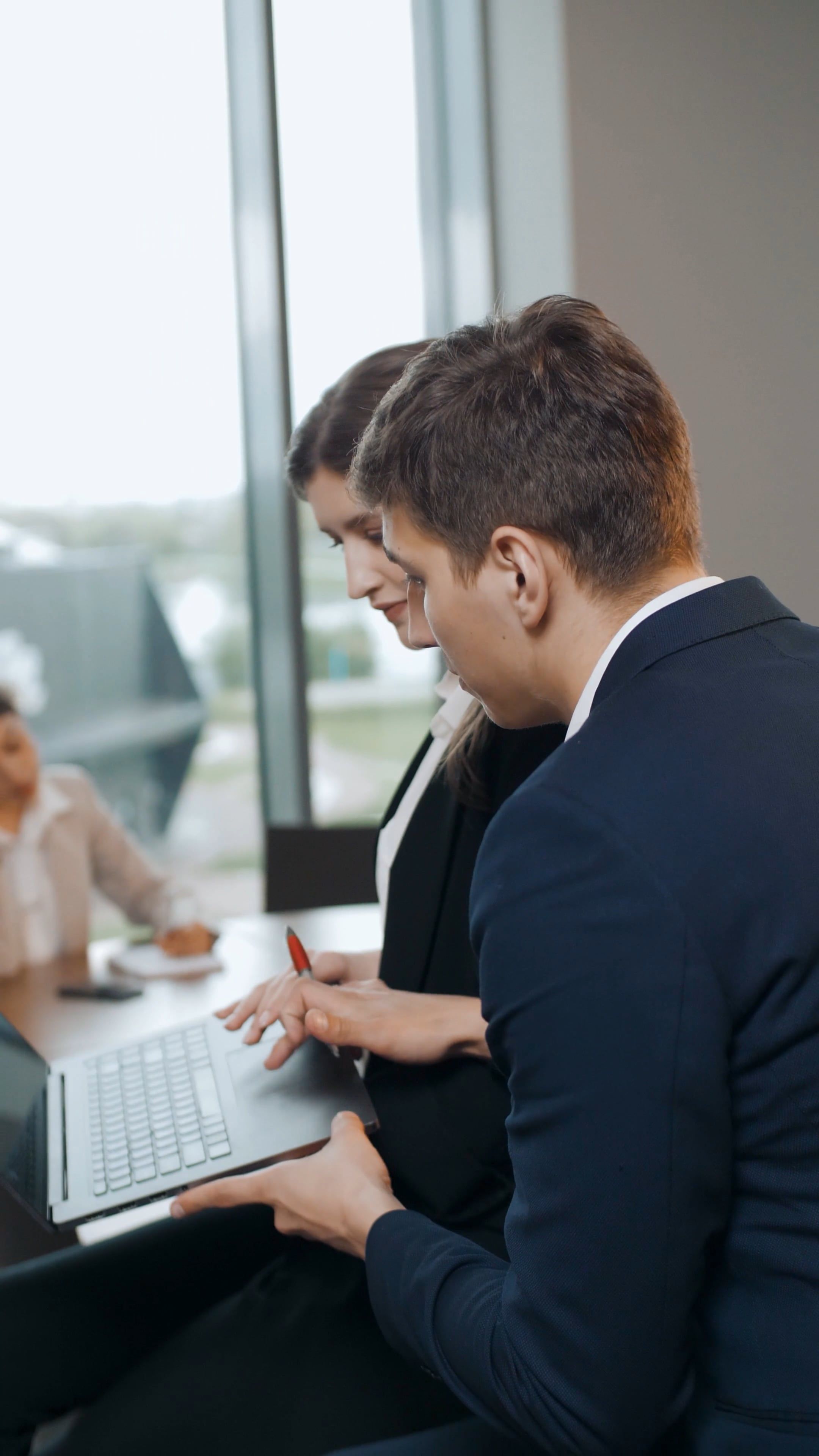Deux professionnels en réunion utilisent un ordinateur portable près d’une grande fenêtre, échanges sérieux. formation économique cse contenu incluse.